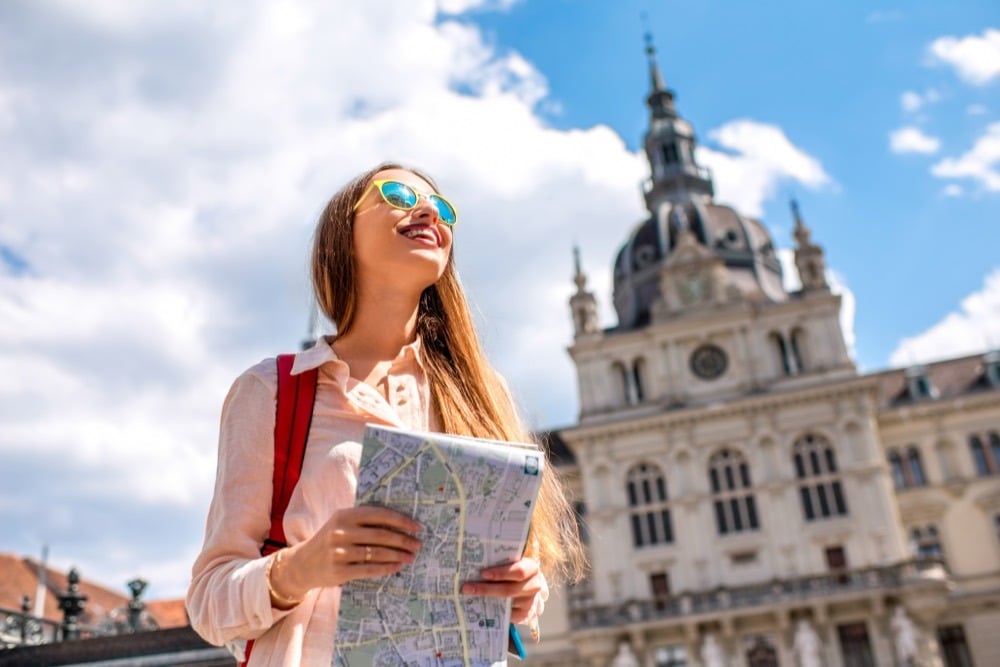 A traveller holding an open map looks up at the domed towers of a historic city building.