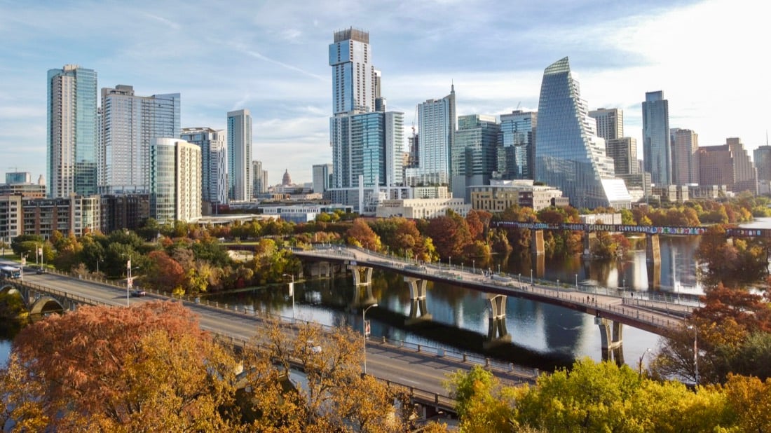 The Austin skyline rising above the green riverbank and a pedestrian bridge.